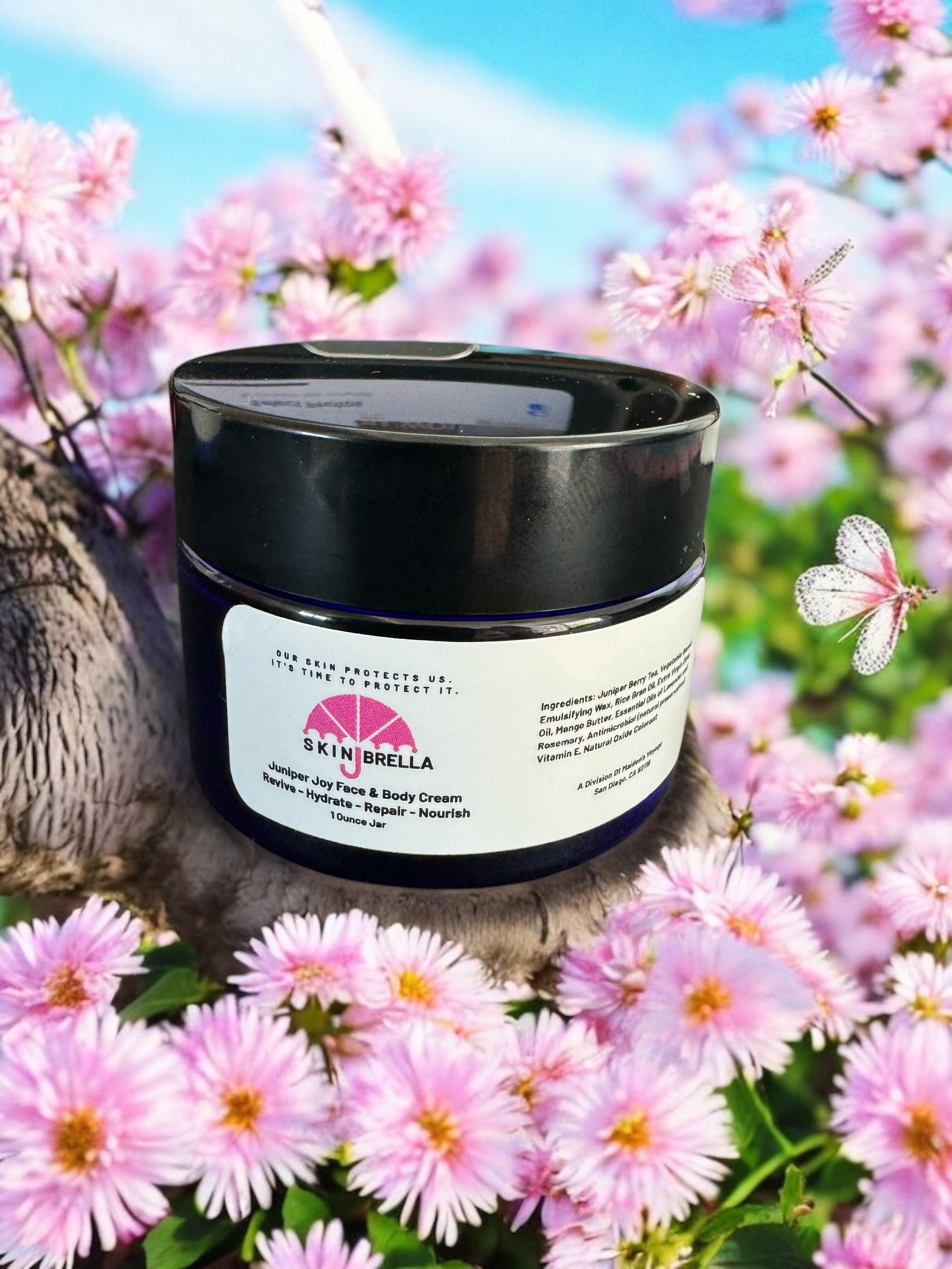 Skin Brella Juniper Joy Face & Body Cream jar surrounded by pink flowers under blue sky